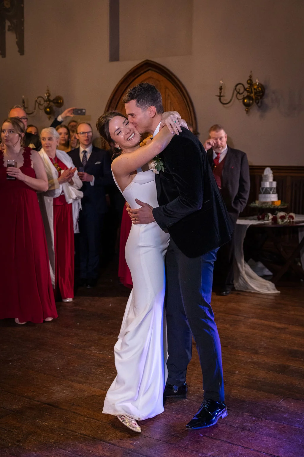 A newlywed couple shares a joyful embrace and a kiss during their first dance on a wooden floor, surrounded by watching guests.
