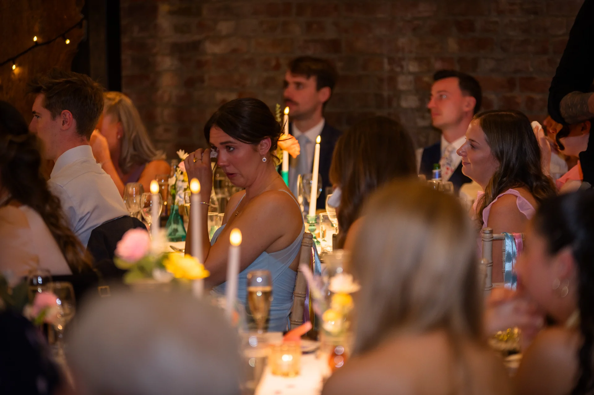 A guest sits at a candlelit wedding reception table against an exposed brick wall, wiping a tear during the speeches.