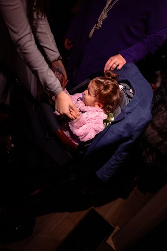 A young toddler in a pink jacket sits comfortably in a pushchair while holding hands with a wedding guest during the evening reception.