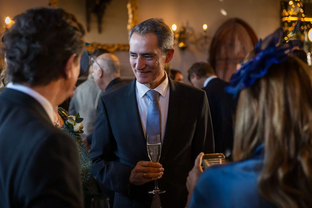 An elegant middle-aged male guest in a charcoal suit and blue tie holds a glass of champagne while mingling with guests at a wedding reception.