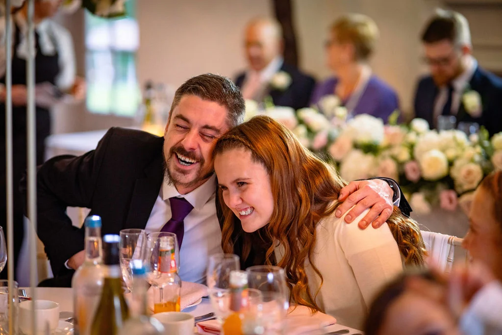 A newly married couple share a joyful, laughing embrace while seated at the top table during wedding speeches, surrounded by guests and floral centrepieces.
