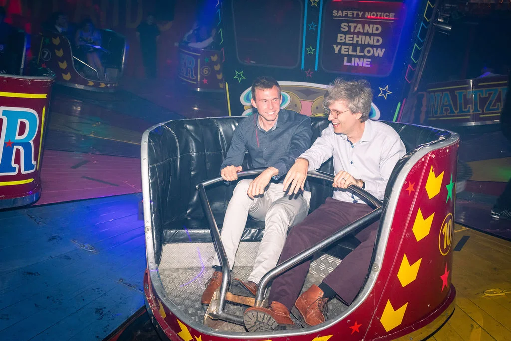 Two grooms laugh joyfully together inside a brightly lit vintage waltzer car at a funfair themed evening wedding reception.