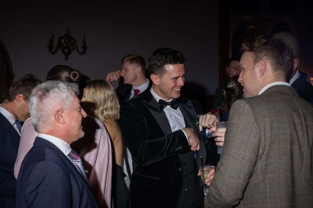 Guests mingling and laughing during an evening wedding reception while holding drinks in a warmly lit room.
