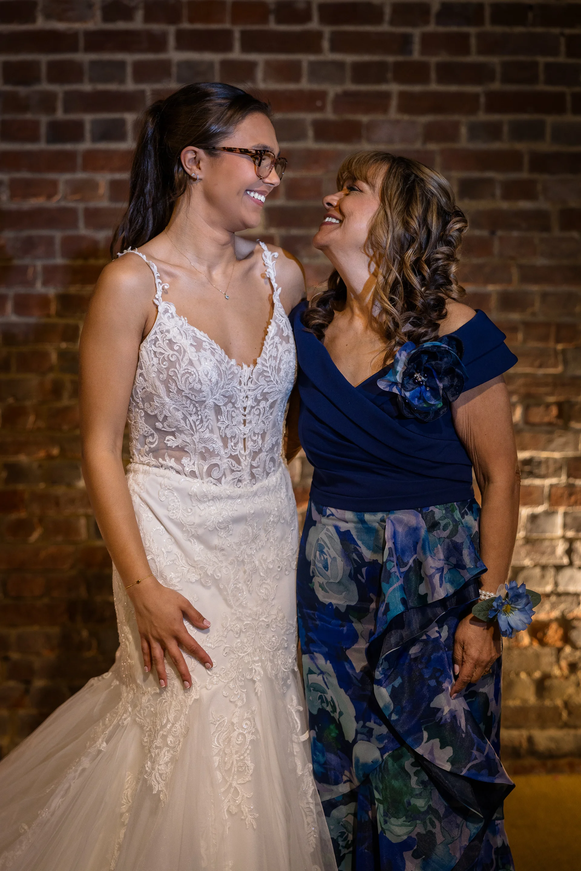A smiling bride wearing a lace gown with exposed boning looks lovingly at her mother, who is dressed in a blue patterned dress against an exposed brick wall.