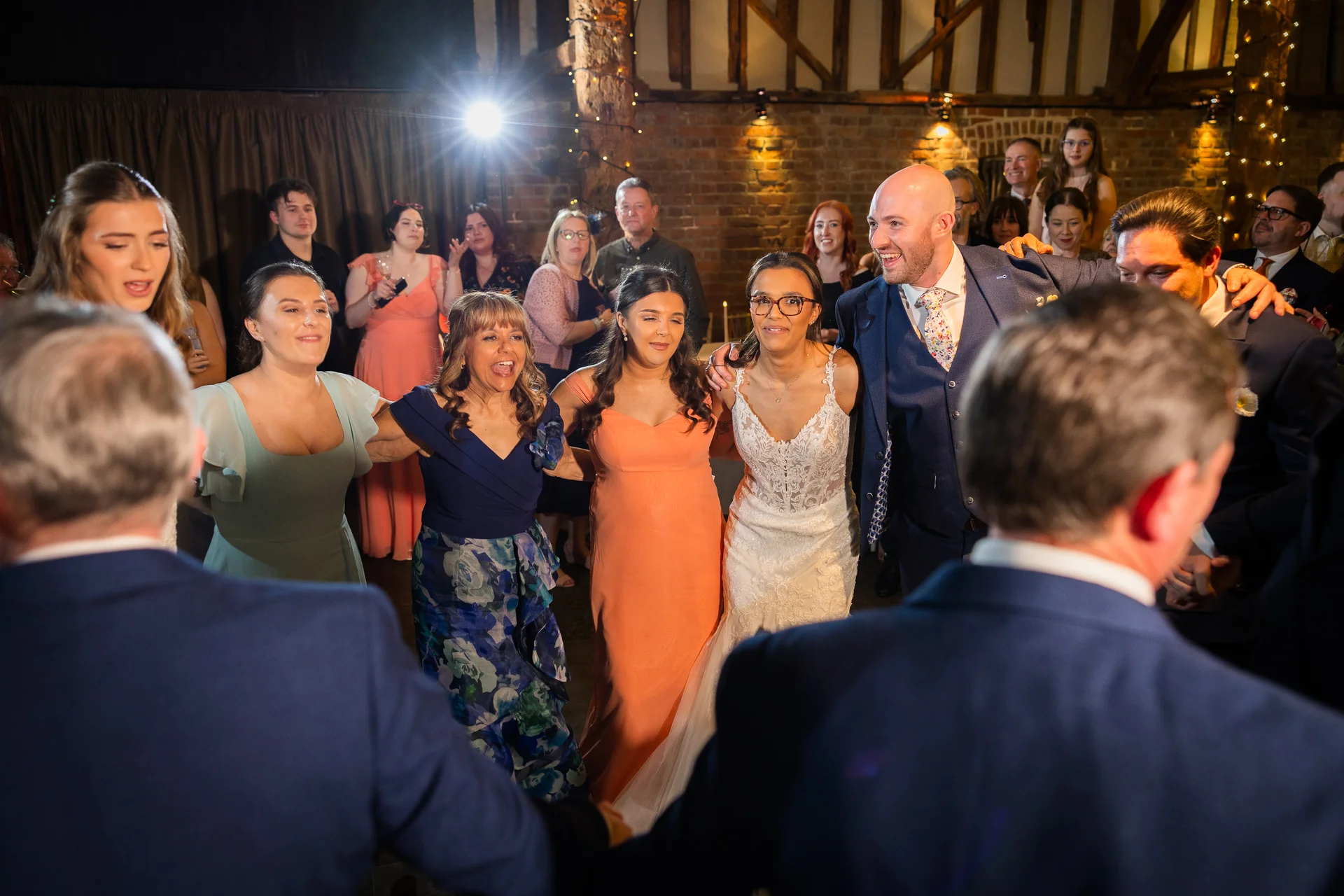 A joyful wedding circle forms on the dance floor inside a rustic barn venue with exposed brick walls and a fairy light canopy.