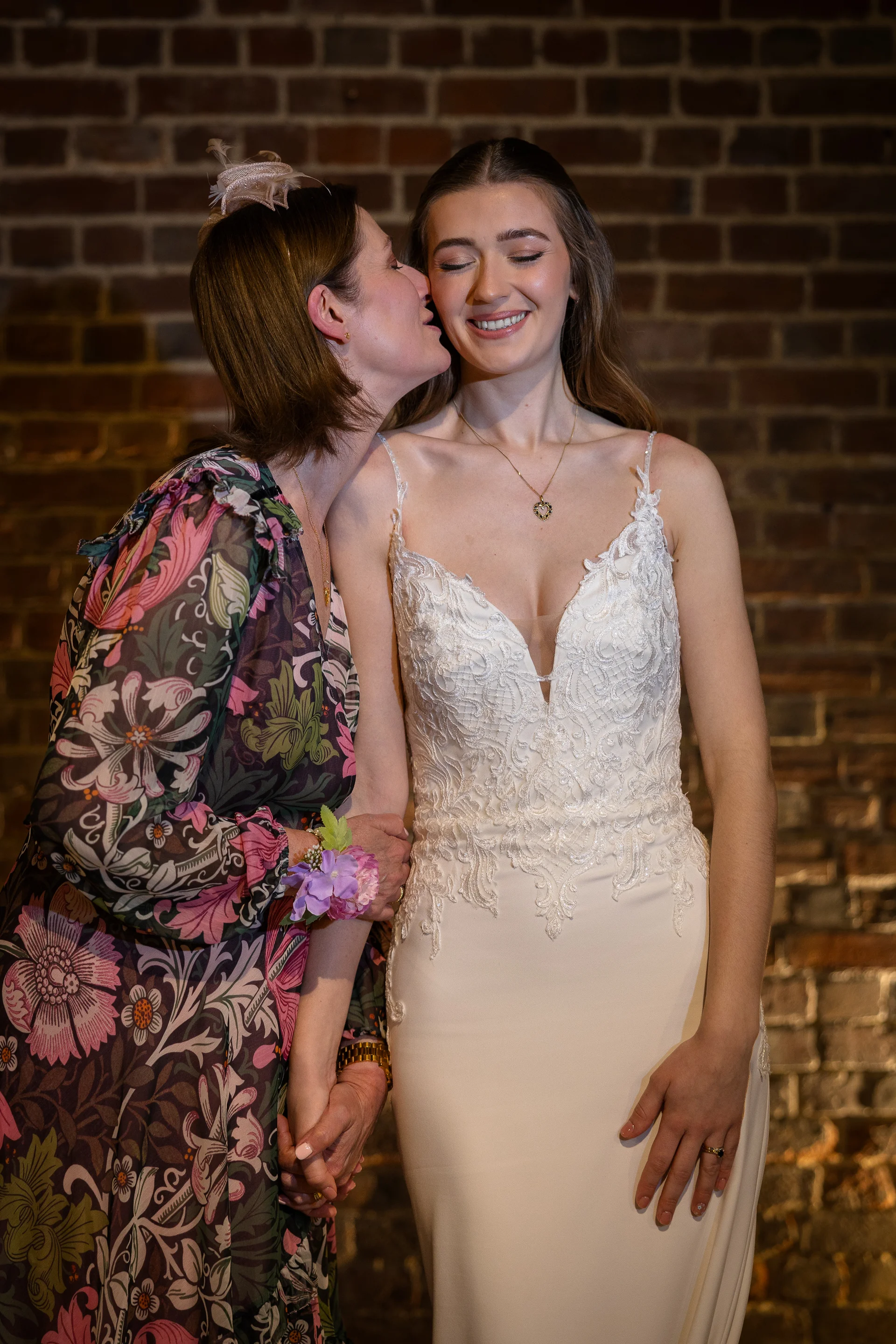 A smiling bride in a lace-detailed slip dress holds hands with her mother, who lovingly kisses her cheek against an exposed brick wall background.