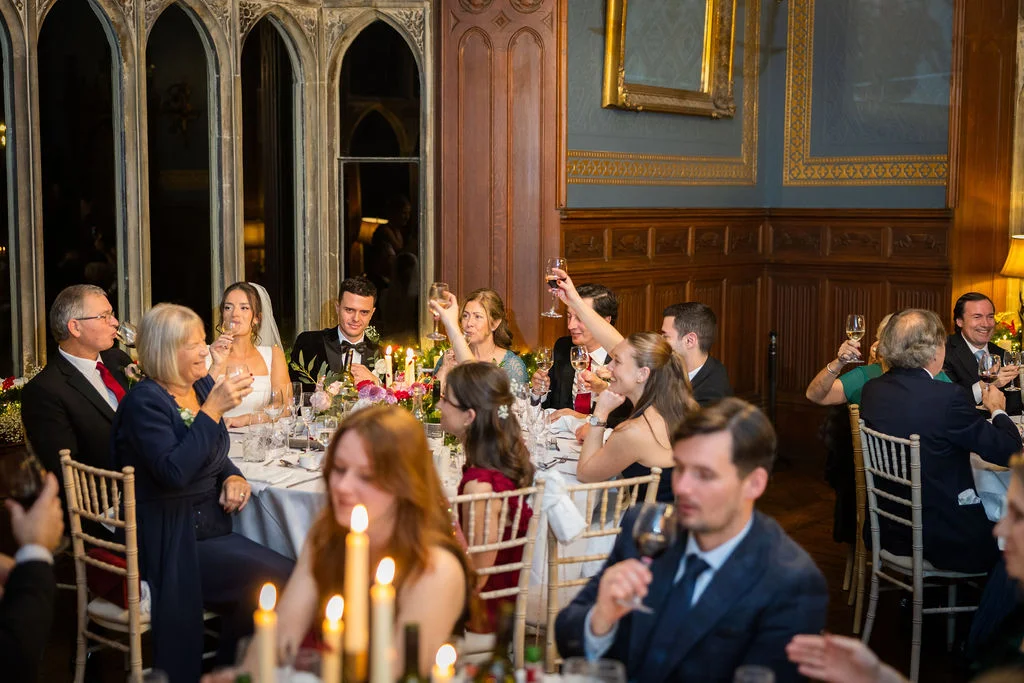 Wedding guests raise their glasses in a toast during the speeches inside a grand wood-panelled hall with gothic windows.