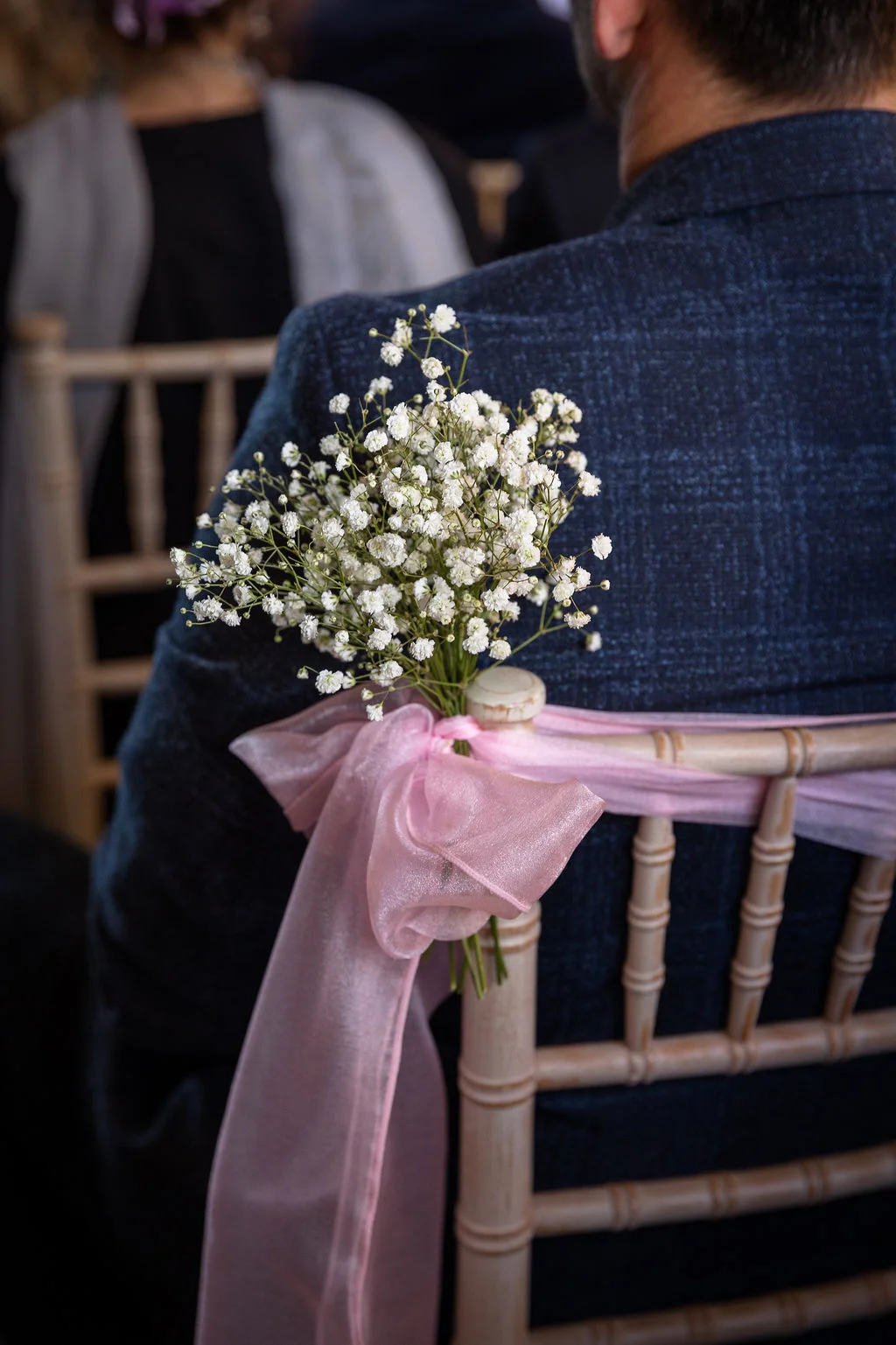 A close-up shot over the shoulder of a seated wedding guest shows a delicate chair decoration featuring a bunch of white baby's breath tied with a pink organza bow.