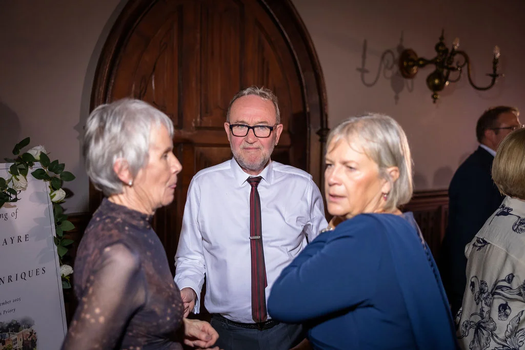 Wedding guests mingle indoors near a wooden arched door and a welcome sign during the evening reception.