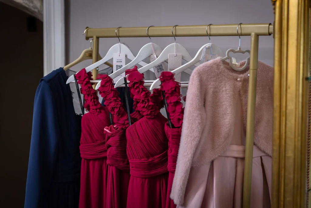 A selection of vibrant pink and burgundy bridesmaid dresses with floral strap details hang neatly on a gold rail during bridal morning preparations.