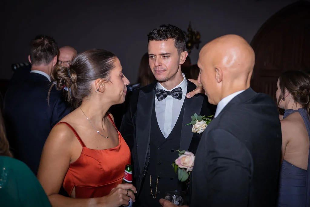 A groom in a velvet tuxedo and bow tie chats with guests at an evening wedding reception while holding a drink.