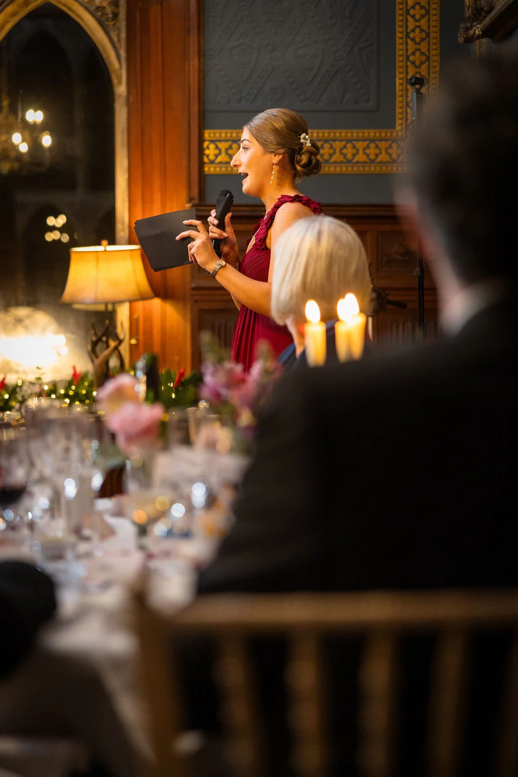 A smiling bride wearing a deep red dress delivers her wedding speech into a microphone, holding notes while standing at a candlelit reception table.