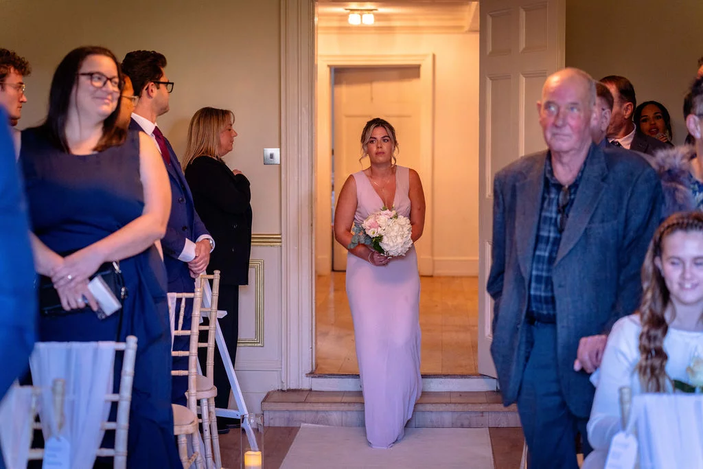 A bridesmaid holding a pink and white floral bouquet enters through an open doorway while wedding guests watch from seated positions.
