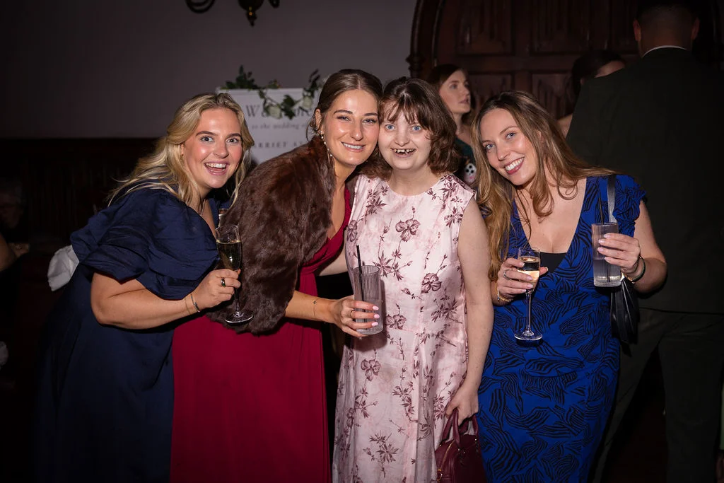 Four smiling female wedding guests hold drinks and pose together during the evening reception, captured using direct flash in a dimly lit indoor venue.