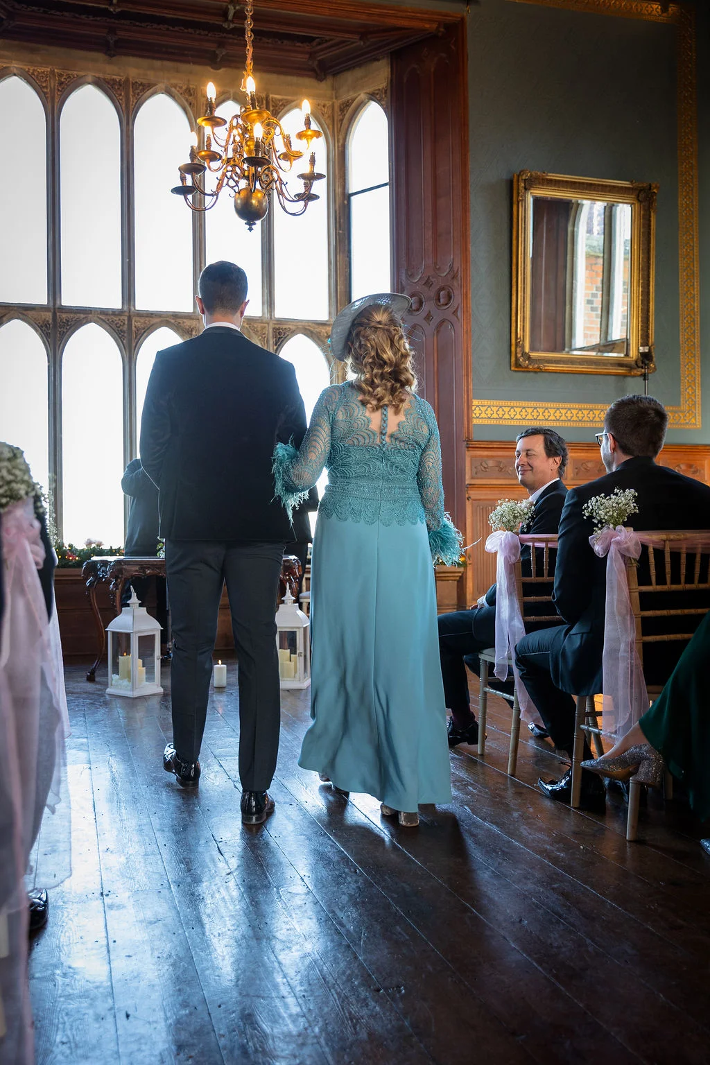 A groom and a mother of the bride walk down the aisle together inside a grand historic venue featuring tall arched windows and wooden flooring.