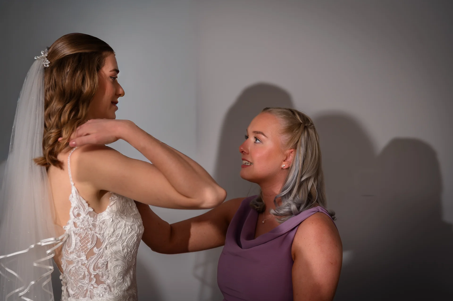 A bridesmaid smiles lovingly at the bride while putting the final touches on her lace wedding gown and veil during morning bridal prep.