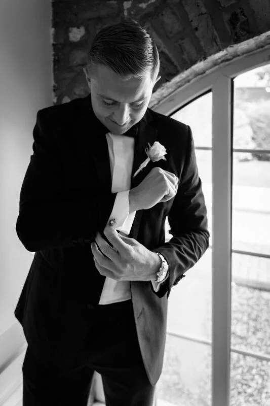 A stylish groom in a black tuxedo adjusts his cufflinks in front of a bright window during his morning preparations.