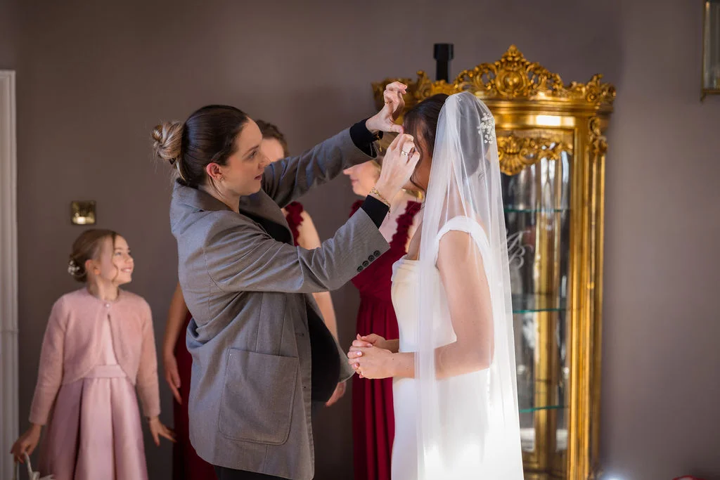 A bride in a white gown and veil has her accessories adjusted by a stylist in a grey blazer during bridal preparations.