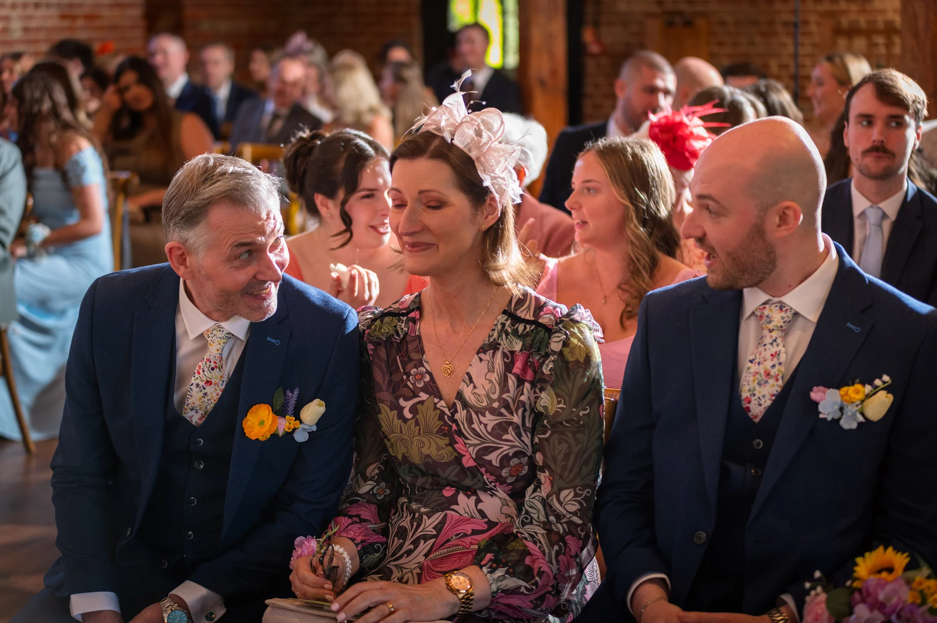 Emotional mother of the bride wearing a floral print dress and fascinator sits between family members during a rustic barn wedding ceremony.