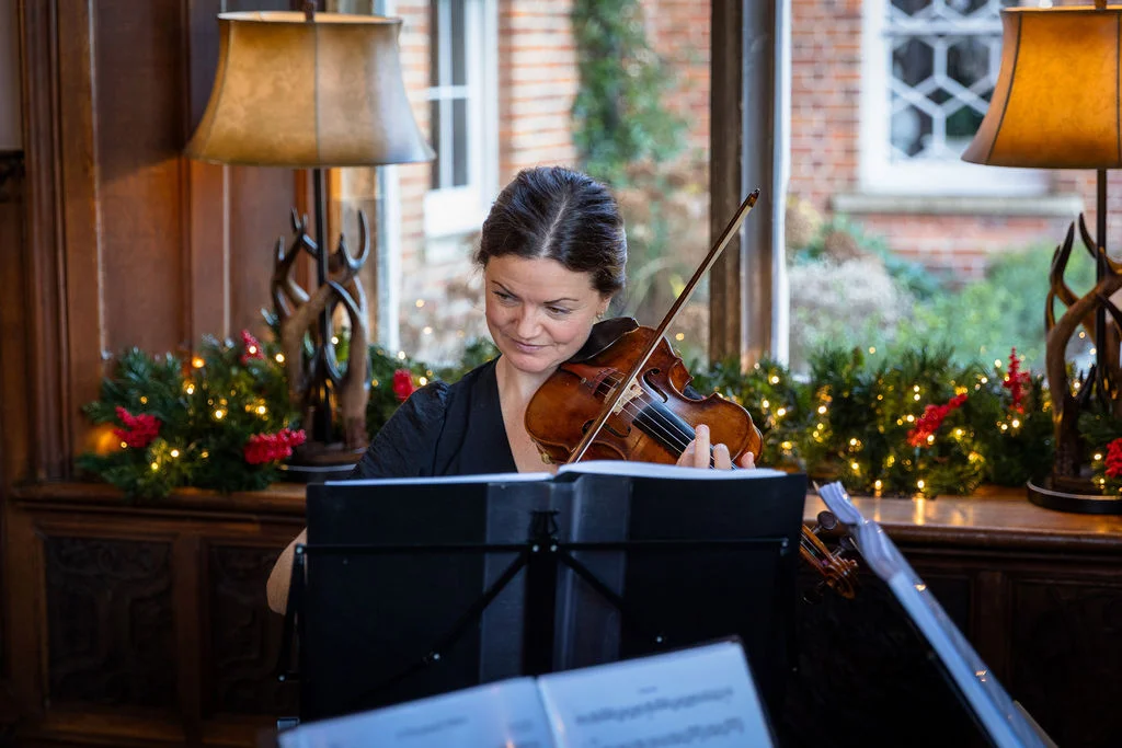 A talented female violinist plays her instrument with intense focus, illuminated by warm window light and festive fairy lights in a wood-panelled room.