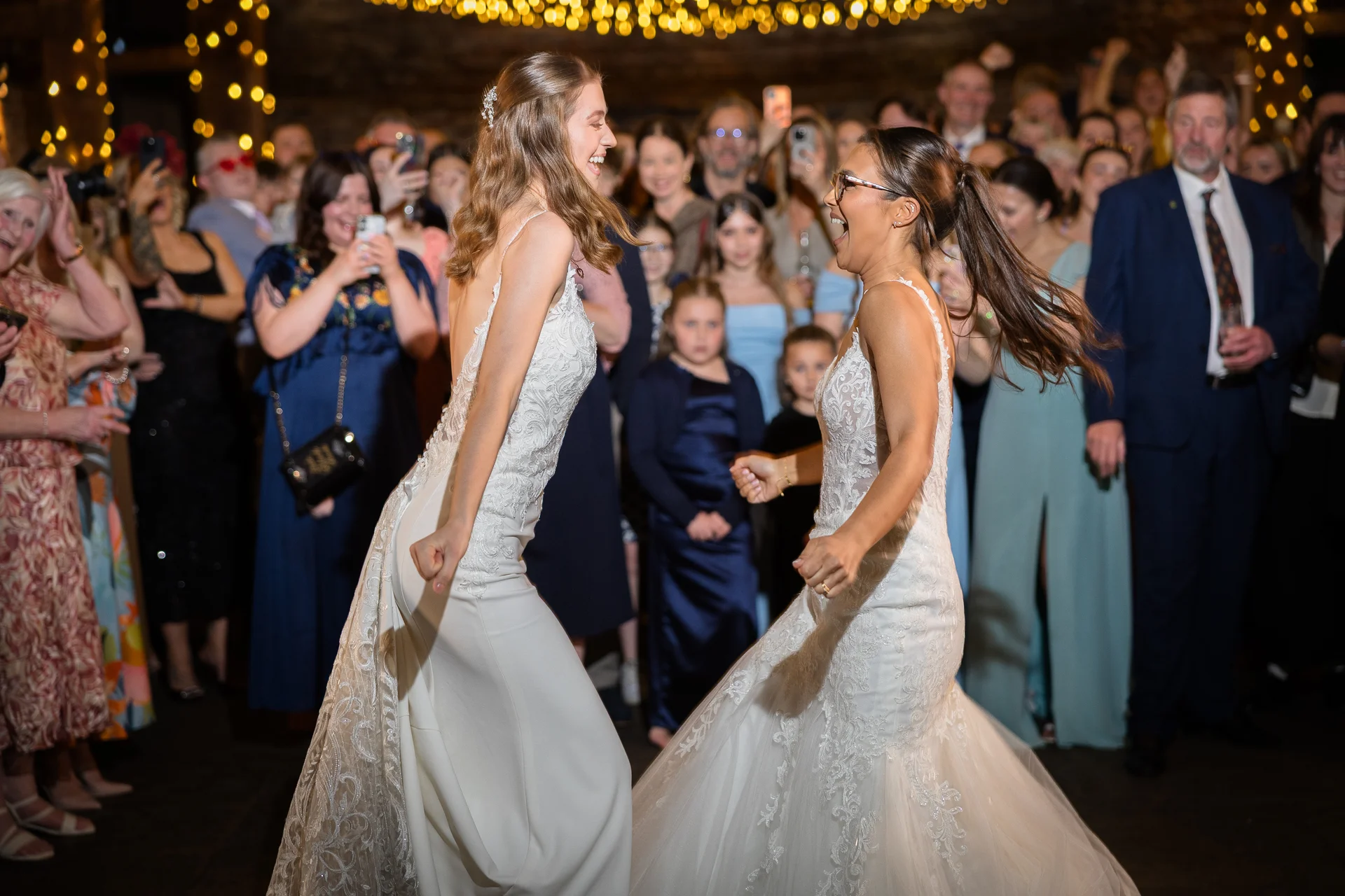 Two smiling brides in lace wedding dresses joyfully dance together on a crowded dance floor beneath glowing festoon lights during their evening reception.