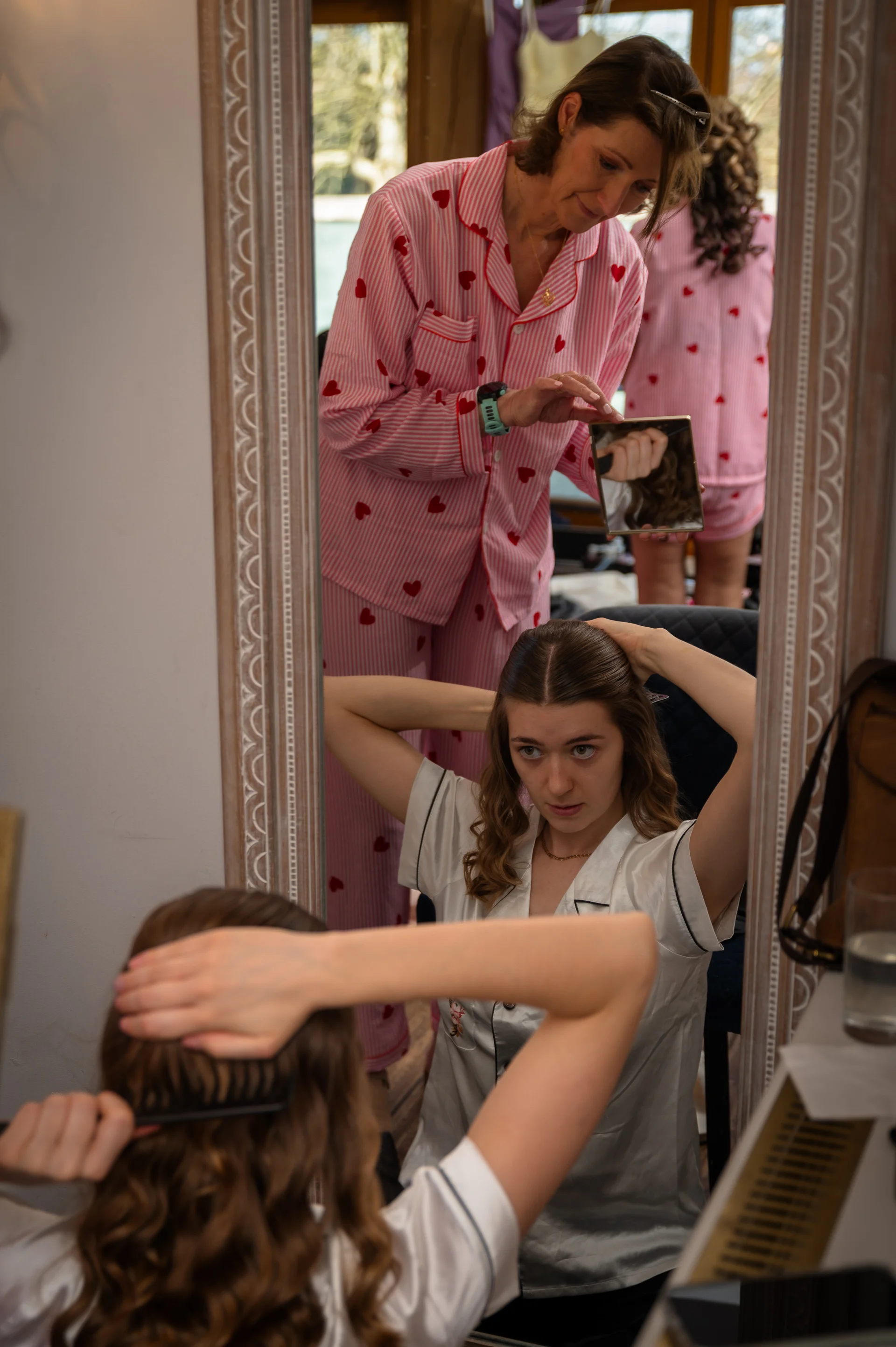 A mother in pink heart-print pyjamas helps style her daughter's hair in front of a large ornate mirror during morning bridal preparations.