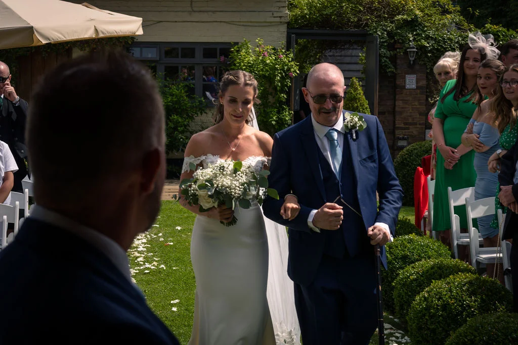 A bride walking down the garden aisle with her father, holding a bouquet and smiling as she approaches the groom.