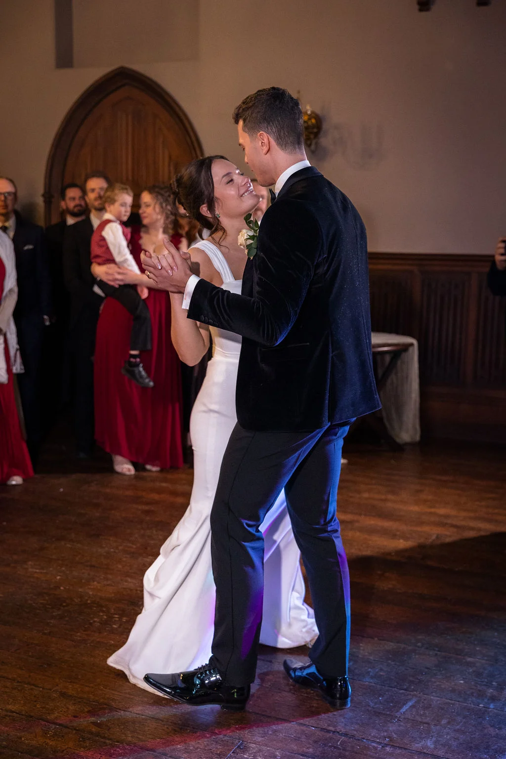 A newlywed couple share a joyful first dance together on a wooden floor indoors, surrounded by watching guests.