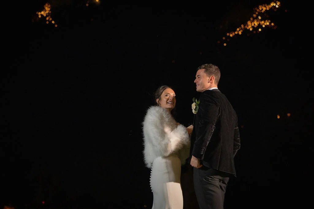 A newlywed couple stands together under a starry night sky during a fireworks display, with the bride wearing a white gown and a fluffy white stole.