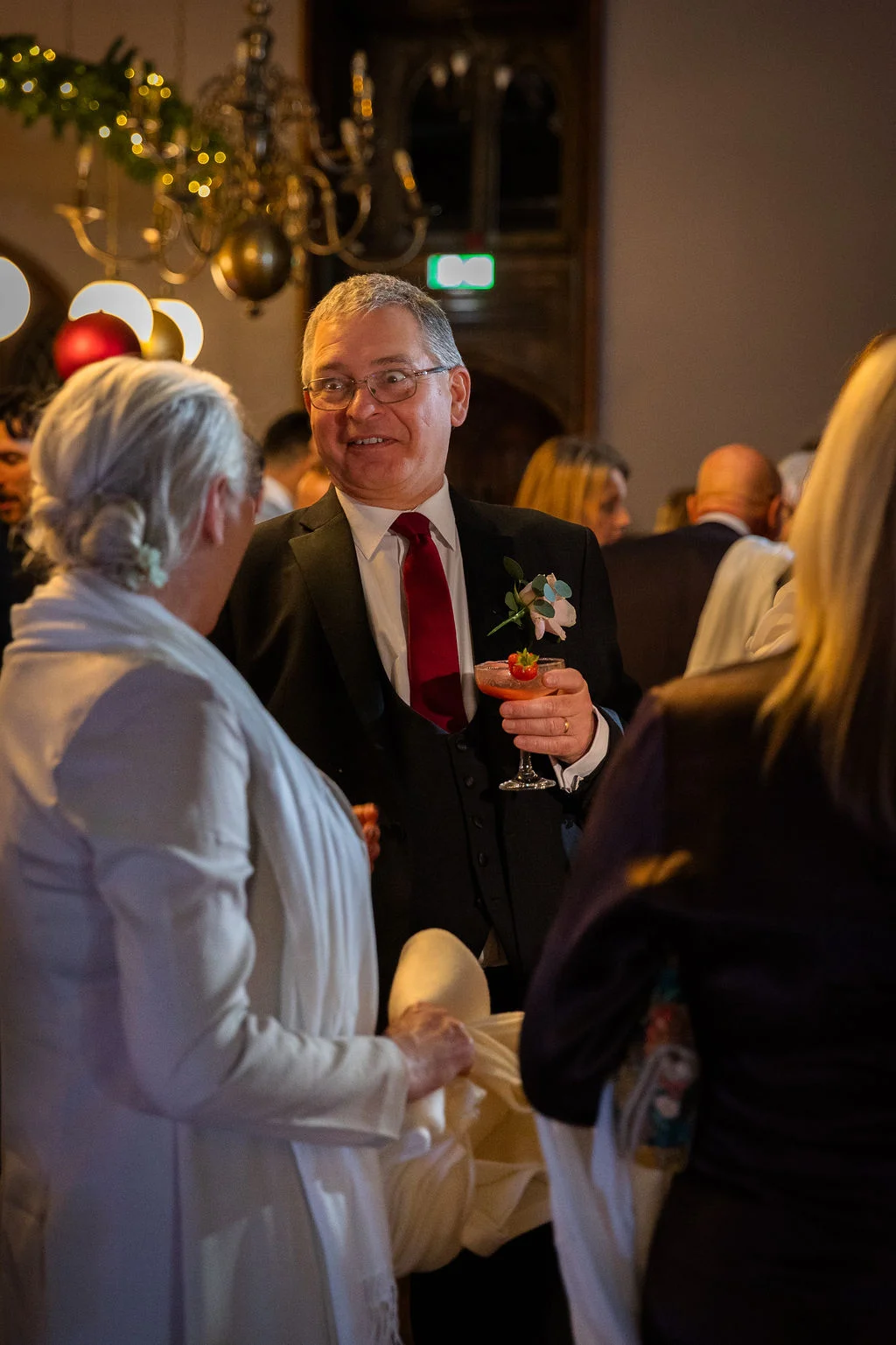 A mature wedding guest in a three piece suit and red tie holds a cocktail while chatting with guests during an evening drinks reception.