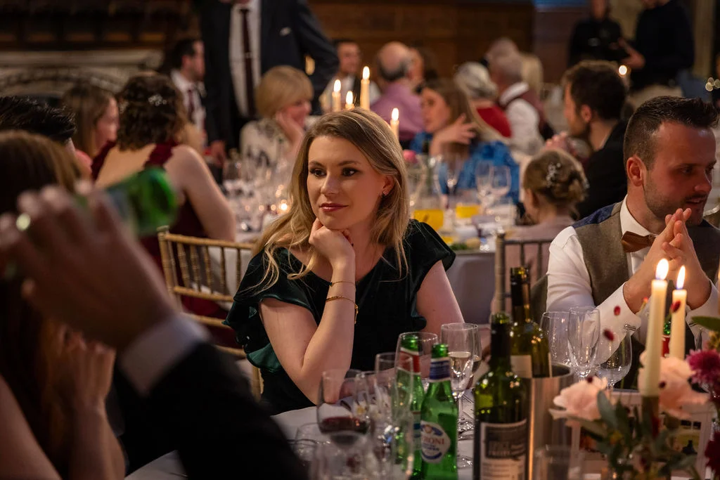 Guests enjoy a candlelit wedding breakfast at long banquet tables, with a woman resting her chin in her hand while looking thoughtfully towards the speaker.