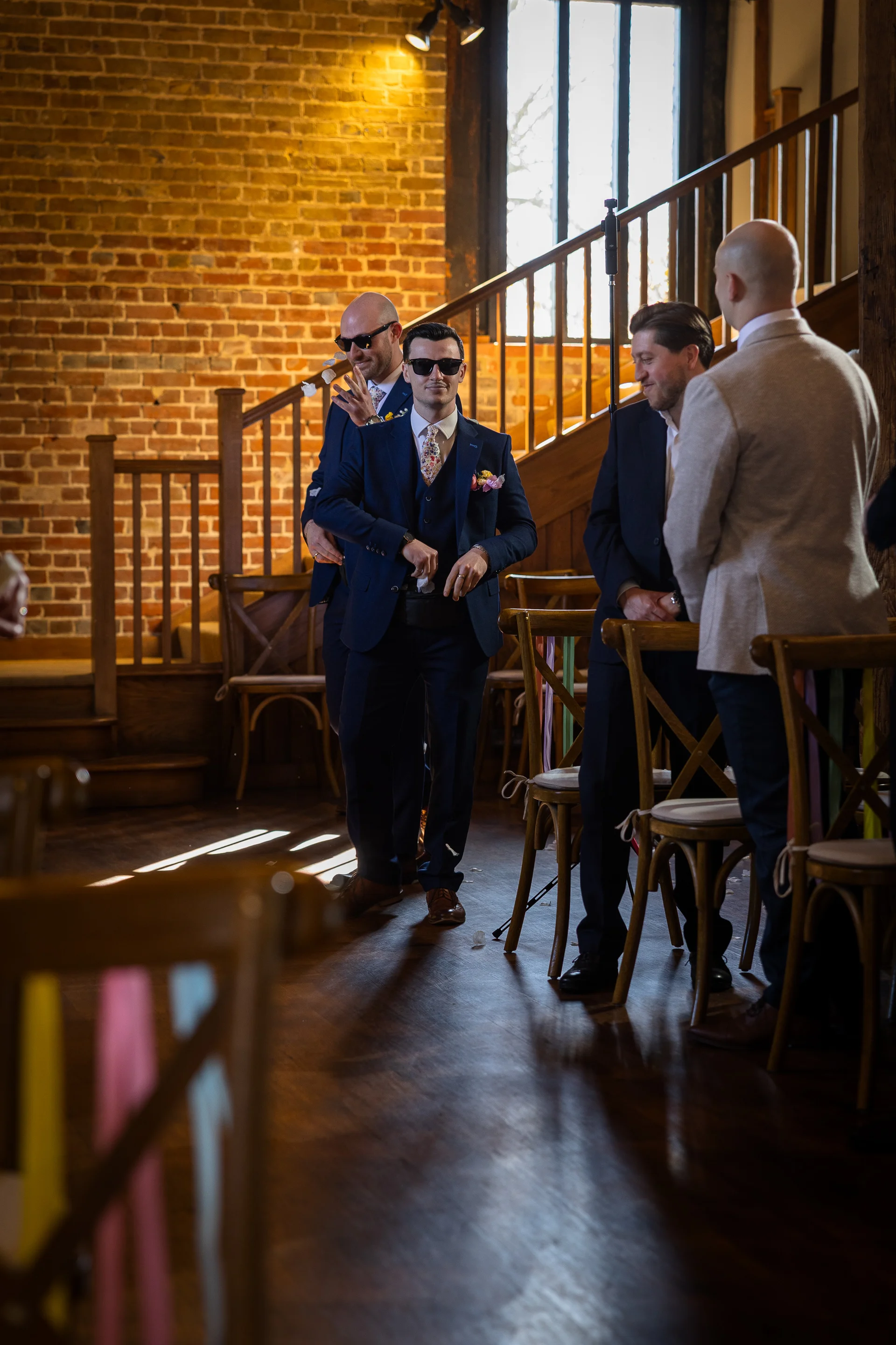 A stylish groom wearing sunglasses and a navy three-piece suit poses with his groomsmen inside a rustic brick barn venue.