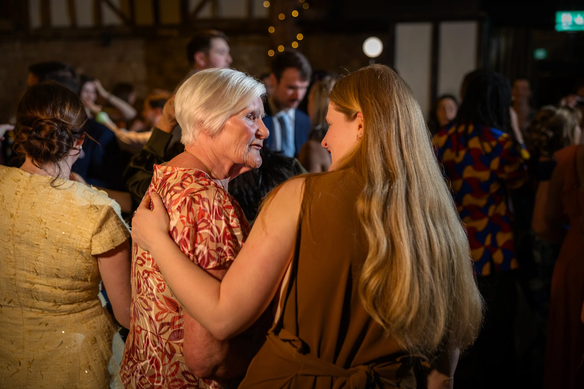 An older woman with white hair shares a tender embrace and conversation with a younger woman with long blonde hair during a rustic evening wedding reception.