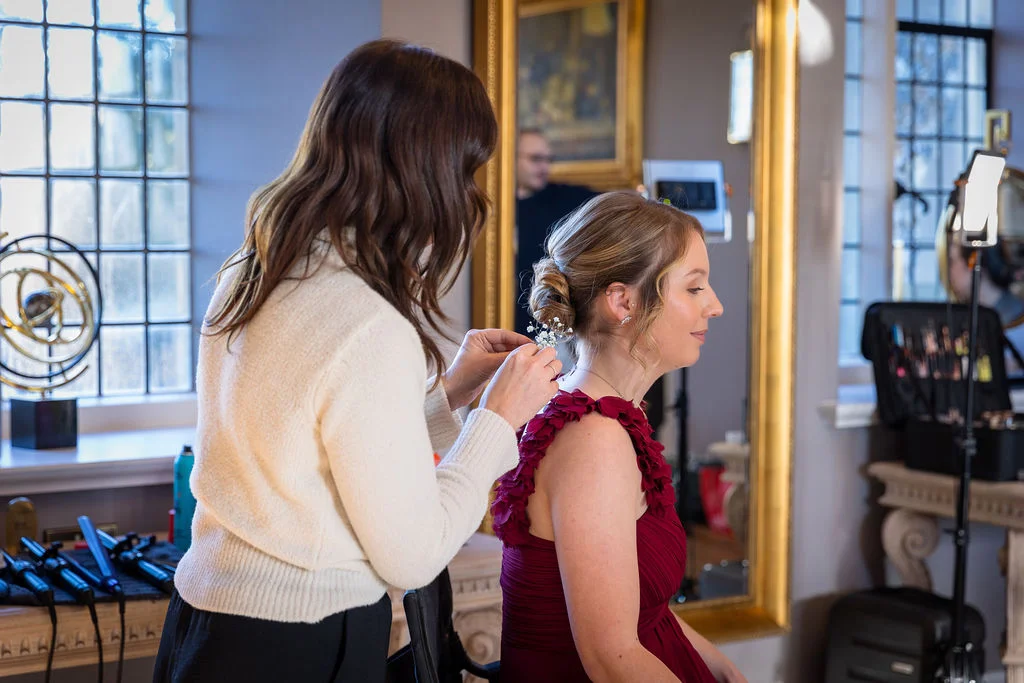 A stylist carefully adds delicate gypsophila flowers into the textured bun of a bridesmaid wearing a burgundy dress during wedding morning preparations.