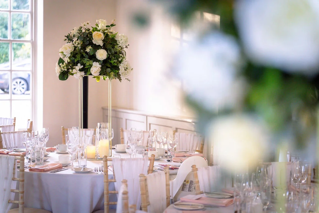 An elegantly dressed round reception table features a tall floral centrepiece of white roses and greenery, soft lighting, and delicate glassware inside a bright venue.