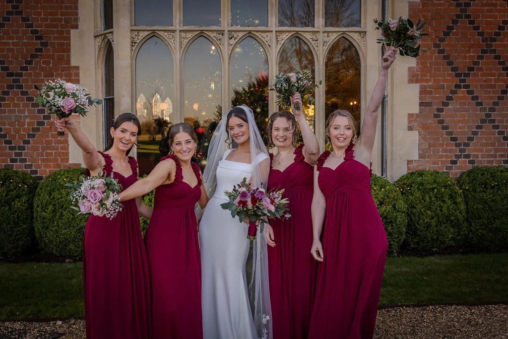 A radiant bride in a white crepe gown and veil poses outdoors with her four bridesmaids wearing matching burgundy dresses and holding bouquets, in front of a historic building with large arched windows.