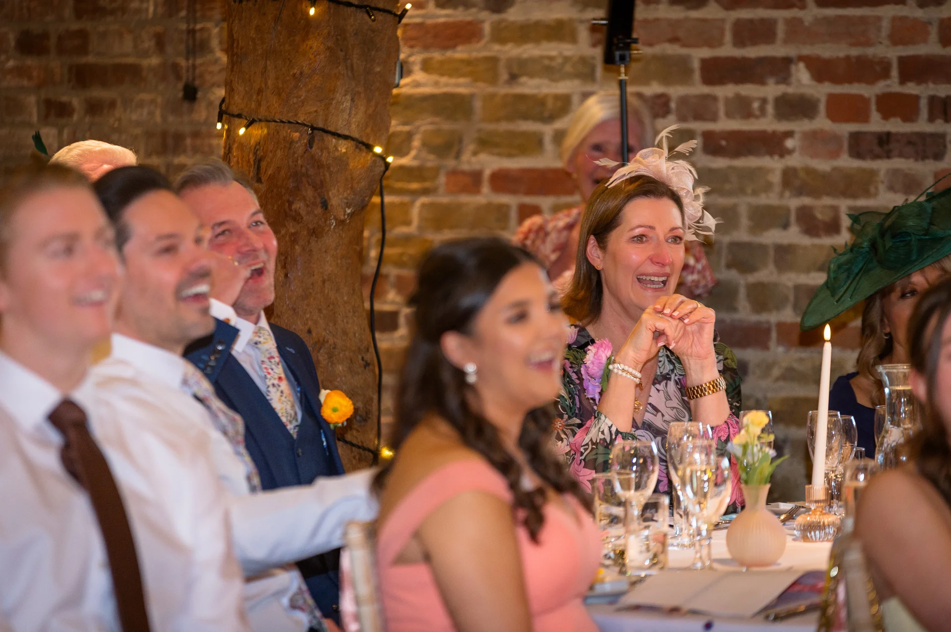 Wedding guests seated at banquet tables inside an exposed brick barn laugh heartily during speeches, with candles glowing on the table.