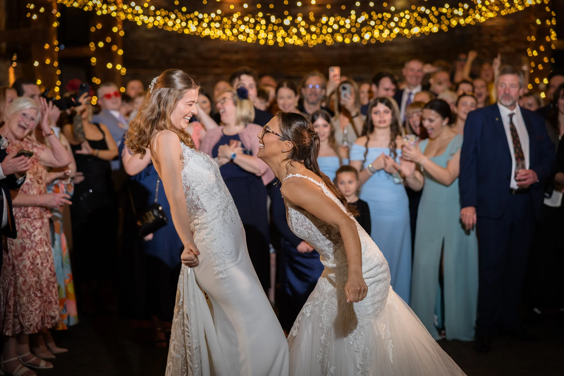 Two smiling brides in white lace wedding dresses lean back playfully while dancing together on a packed dance floor beneath a canopy of twinkling fairy lights.