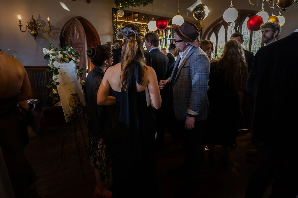 Wedding guests mingle indoors near a welcome sign and bar decorated with festive baubles and soft lighting.