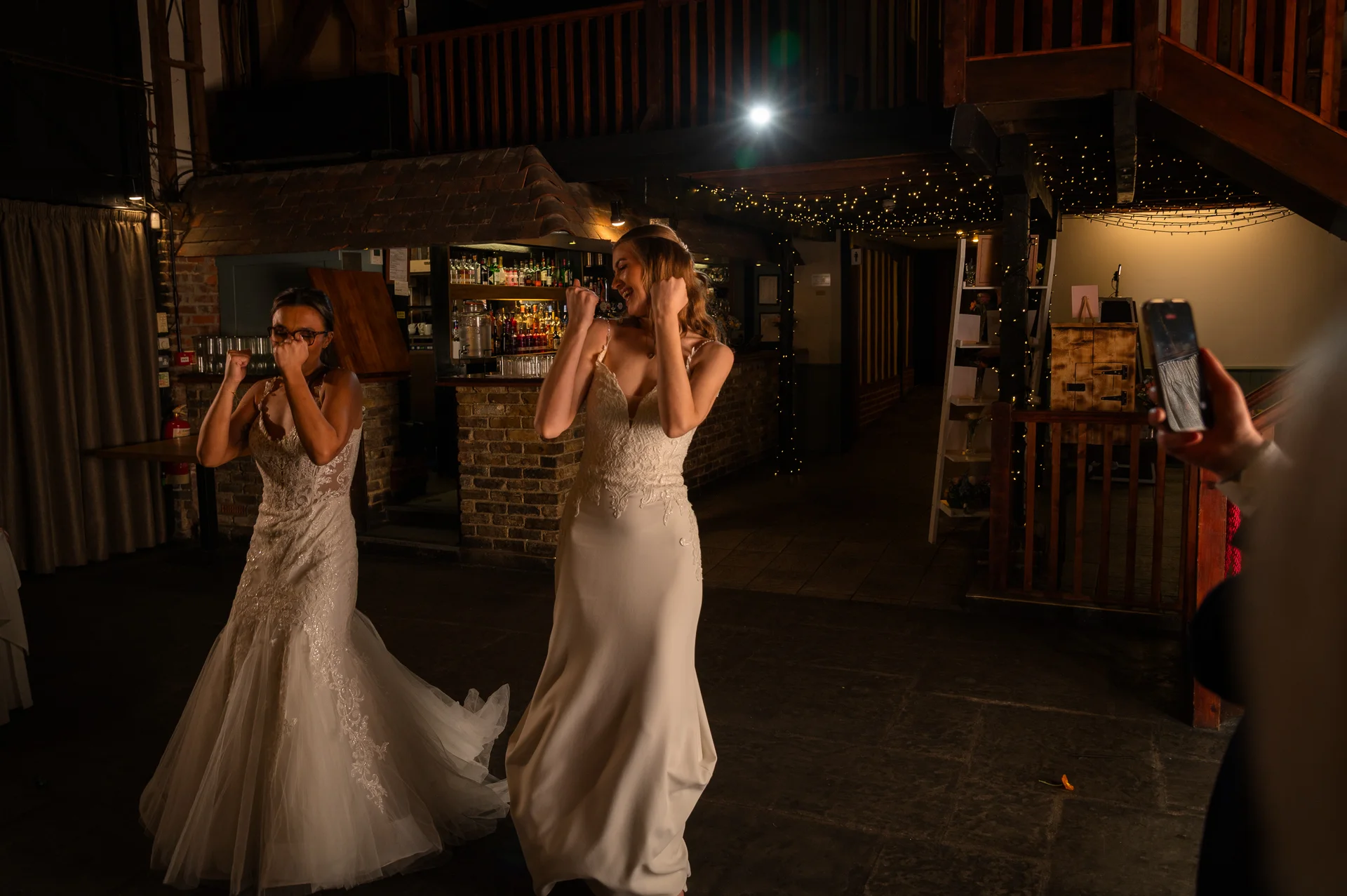 Two brides in white wedding dresses joyfully celebrate and dance together indoors at night under fairy lights in a rustic barn venue.
