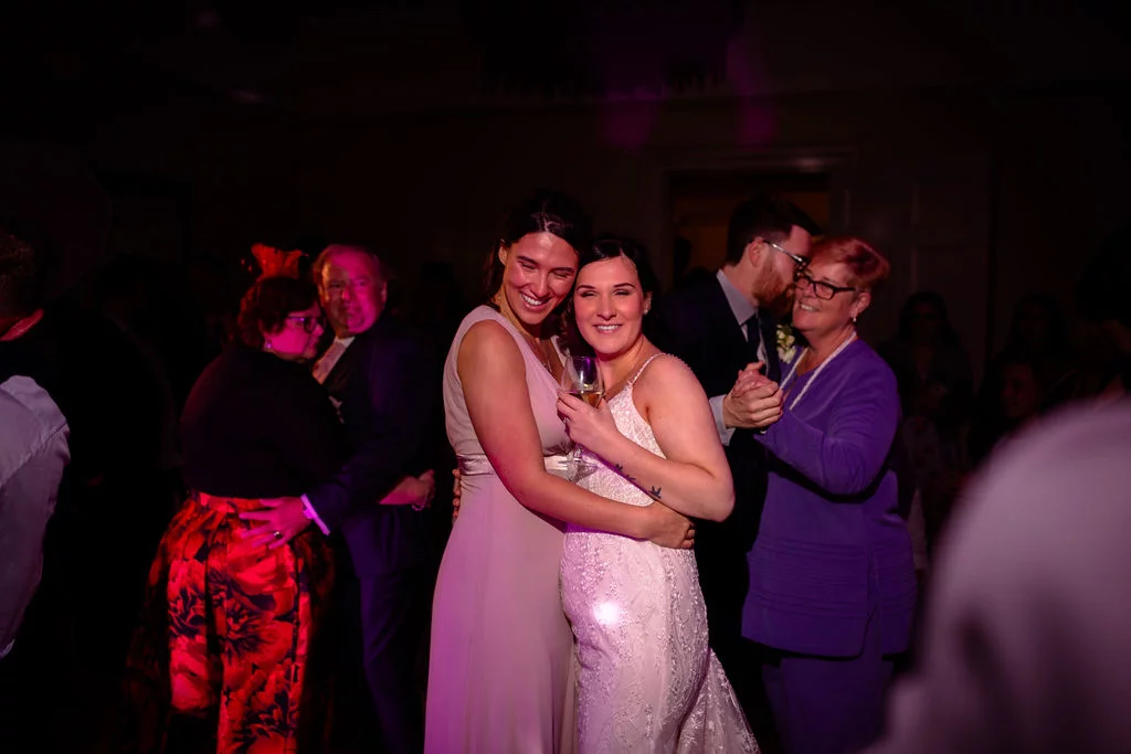 Happy guests embrace and dance together on a lively evening reception dance floor illuminated by vibrant pink and purple lighting.