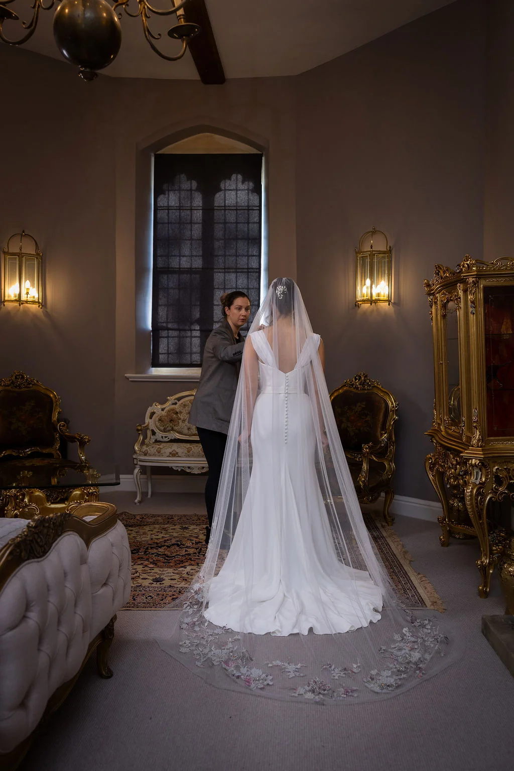 A bride stands facing away in an elegant manor house room as a stylist makes final adjustments to her long embroidered veil and sleek crepe wedding dress.