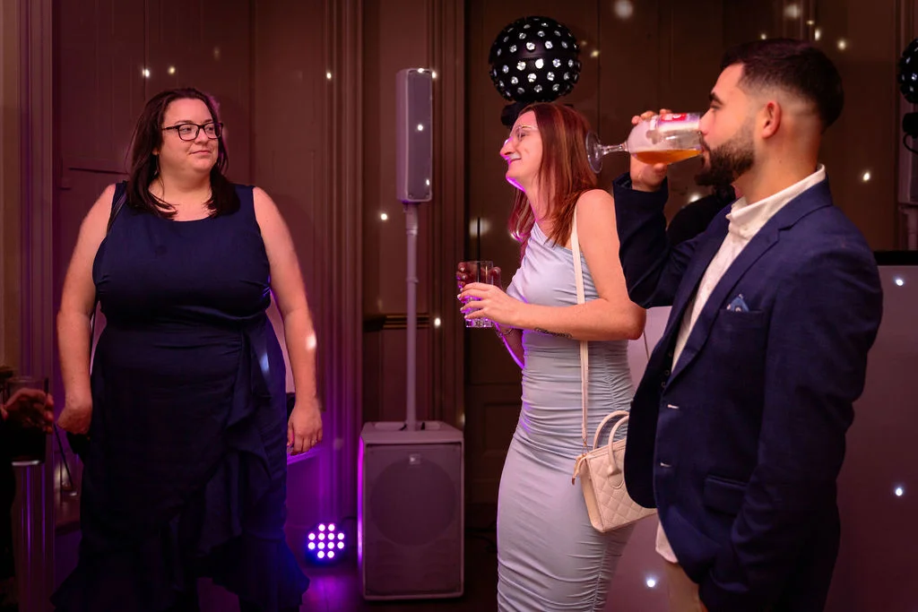 Wedding guests enjoy drinks and conversation during the evening reception, with a man drinking from a glass and women chatting nearby under party lighting.