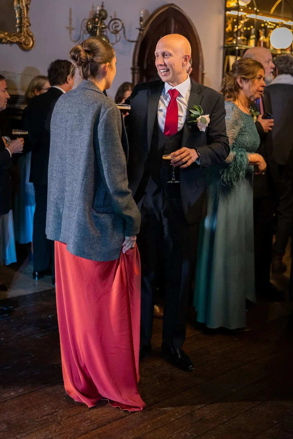 A guest in a three-piece suit and bright red tie laughs while mingling and holding a cocktail during a wedding reception.