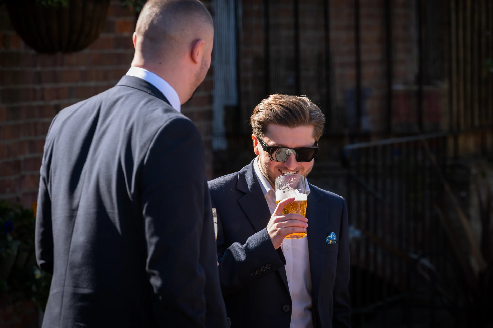 A guest in a navy suit and sunglasses smiling while holding a pint glass of beer outdoors during a wedding drinks reception.