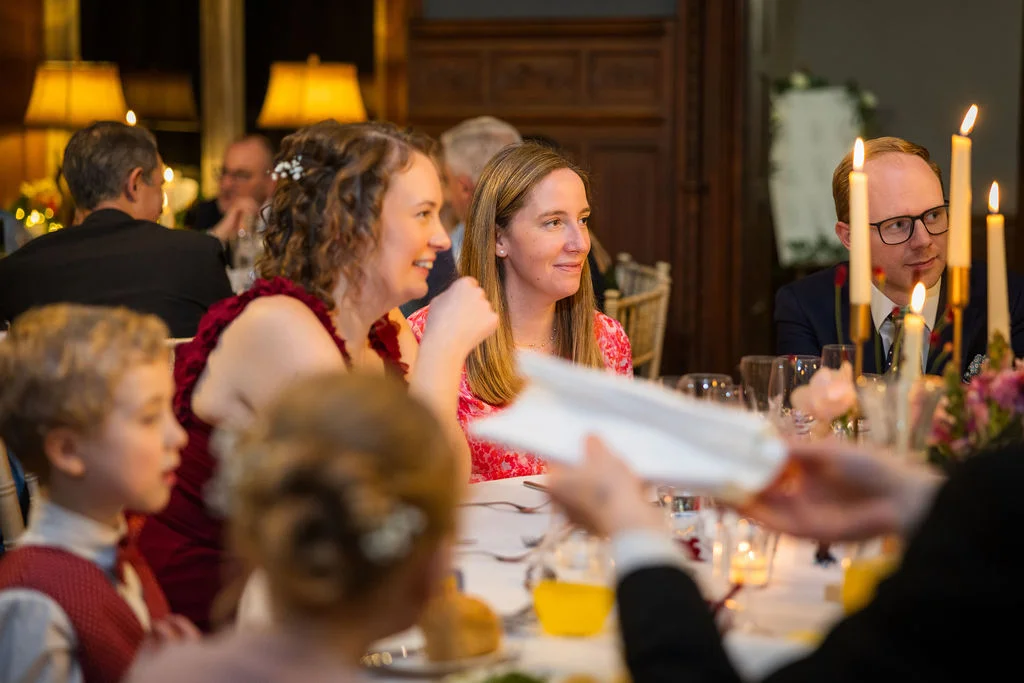 Wedding guests smiling and listening during the wedding breakfast speeches in a warmly lit, wood-panelled reception room.