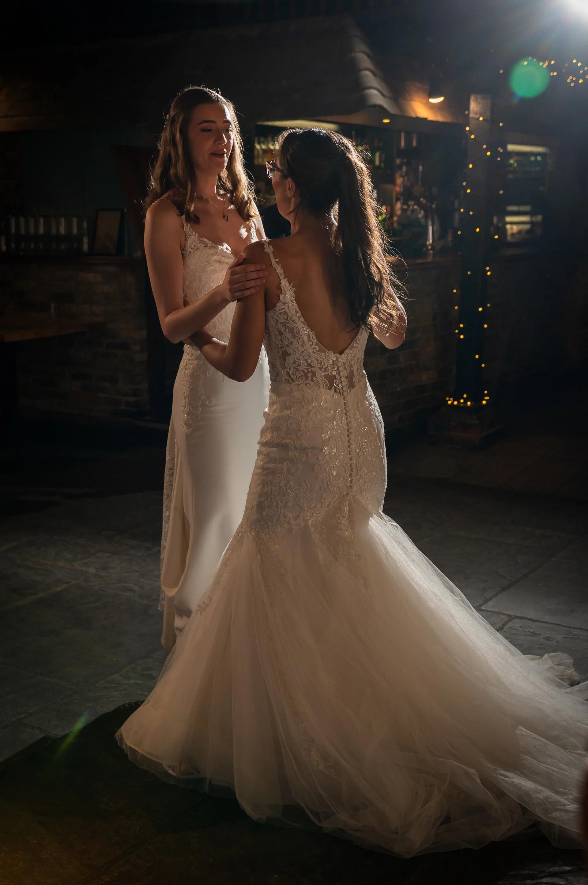 Two brides share a tender moment dancing together in a dimly lit pub venue with exposed brick walls and warm fairy lights.