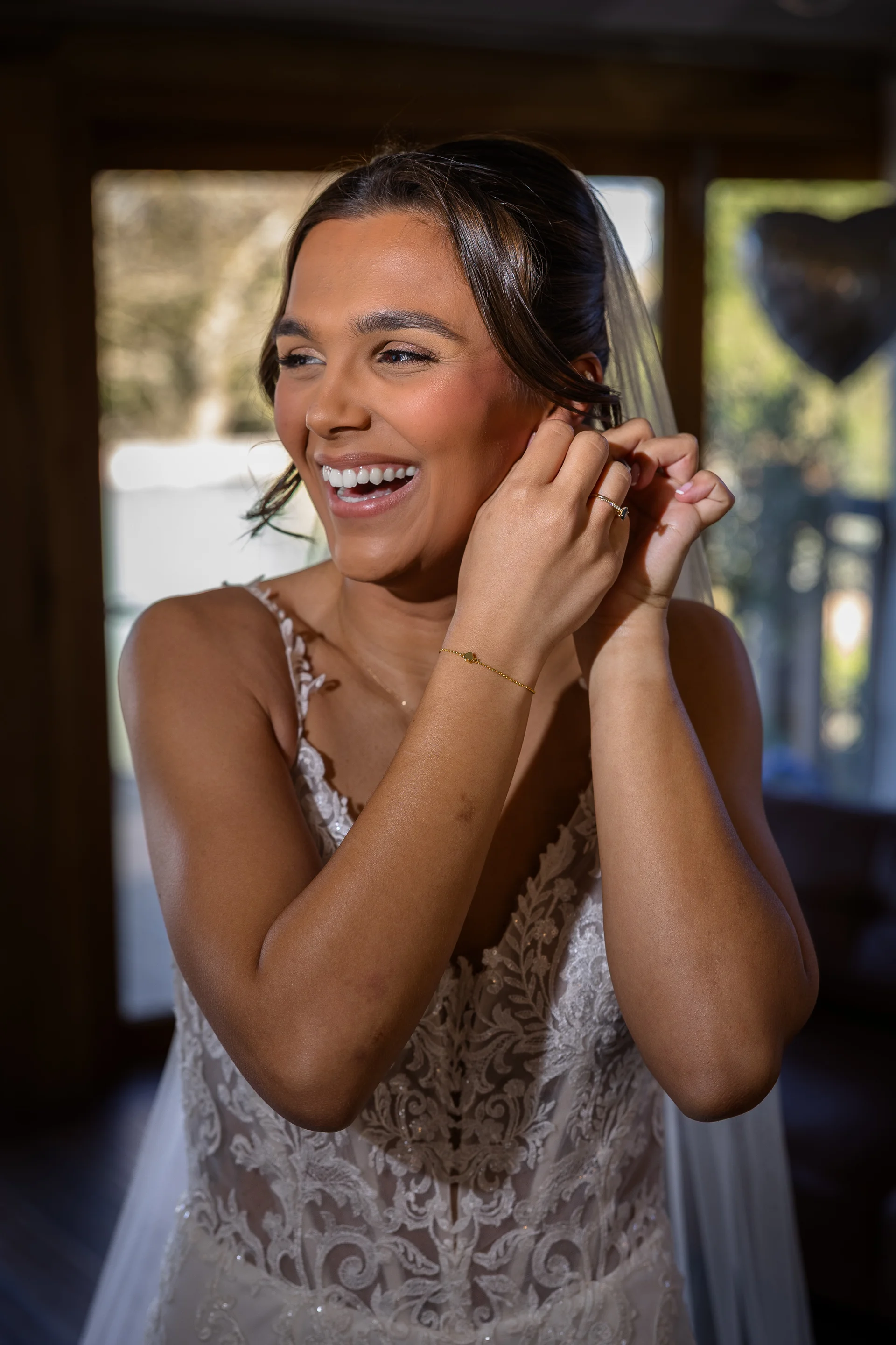 A joyful bride wearing a lace wedding dress with spaghetti straps smiles brightly while putting on her earrings during morning preparations.