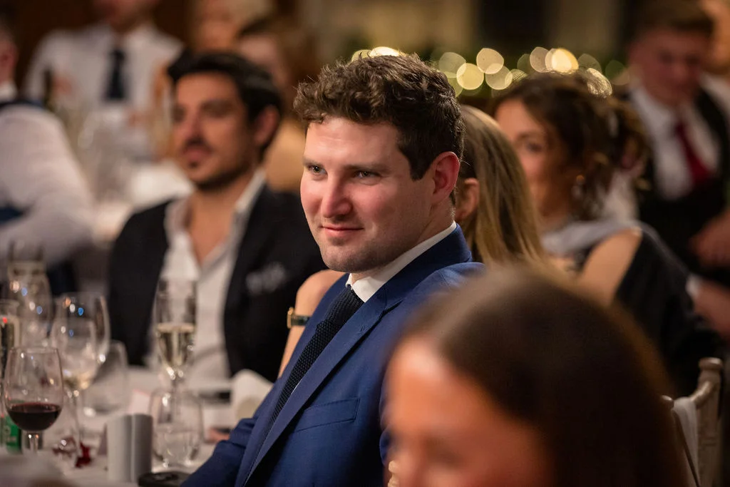 A wedding guest in a blue suit smiles attentively while seated at a crowded reception table during the speeches.
