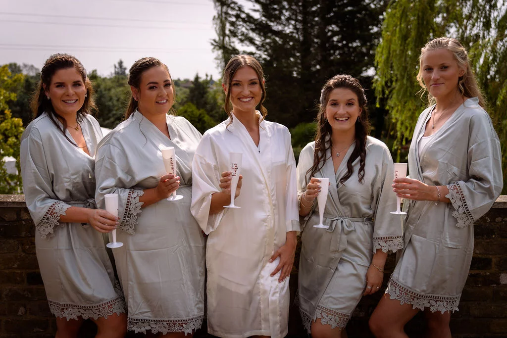 A smiling bride in a white robe and four bridesmaids in matching sage green lace-trimmed robes hold champagne flutes outdoors during morning preparations.